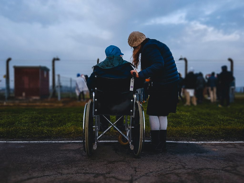 A woman stands beside a woman in a wheelchair, looking away from the camera at a fence with many other people out of focus.