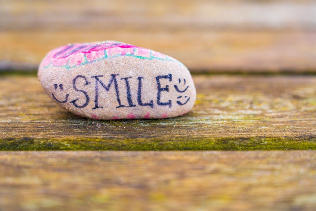 A rock sits on a park bench up close. The rock has many different colours and words scribbled on it. Smile faces us directly with 3 smiling faces drawn to the side of the word