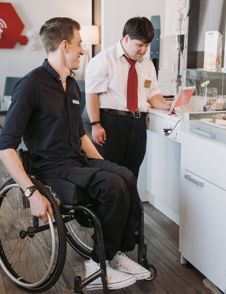A man in a wheelchair and a second standing man look towards a notebook. They are in a working environment