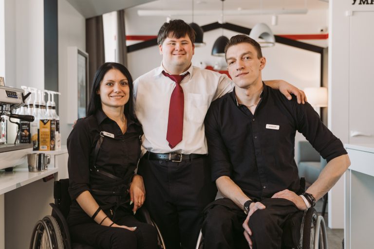 Three people look towards the camera with smiles, two people sit in wheel chairs while the middle man stands up.